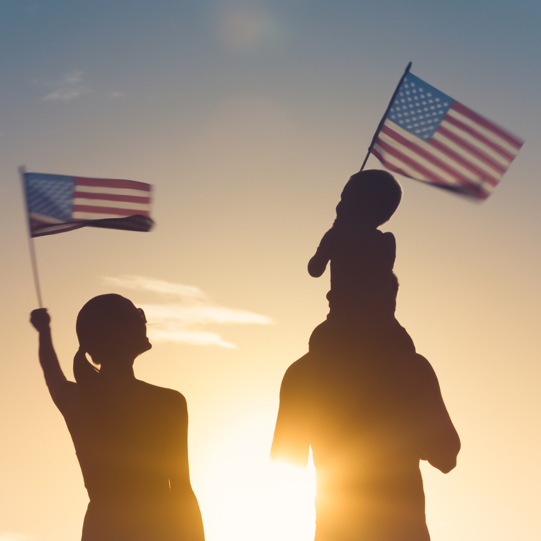 Two silhouettes walking towards a sunset with children on their shoulders holding American Flags. 